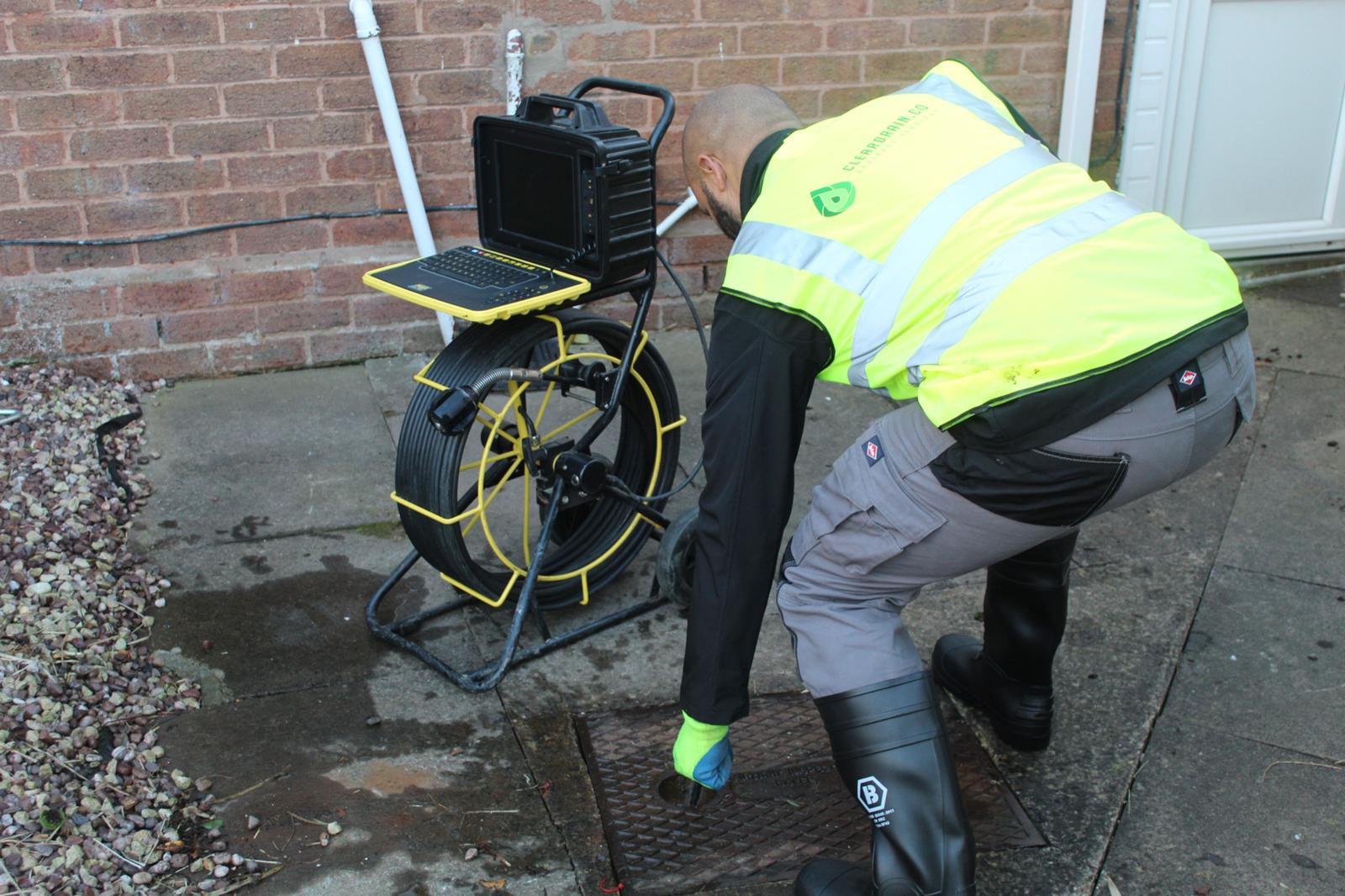 A Clear Drain engineer in branded hi-vis lifting a manhole cover, with the CCTV camera reel and control unit set up alongside