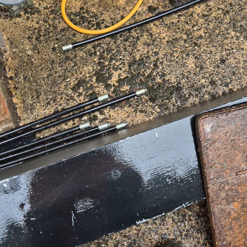 Drain rods laid out on wet paving beside a lifted cast-iron manhole cover, with a jetting hose coiled alongside
